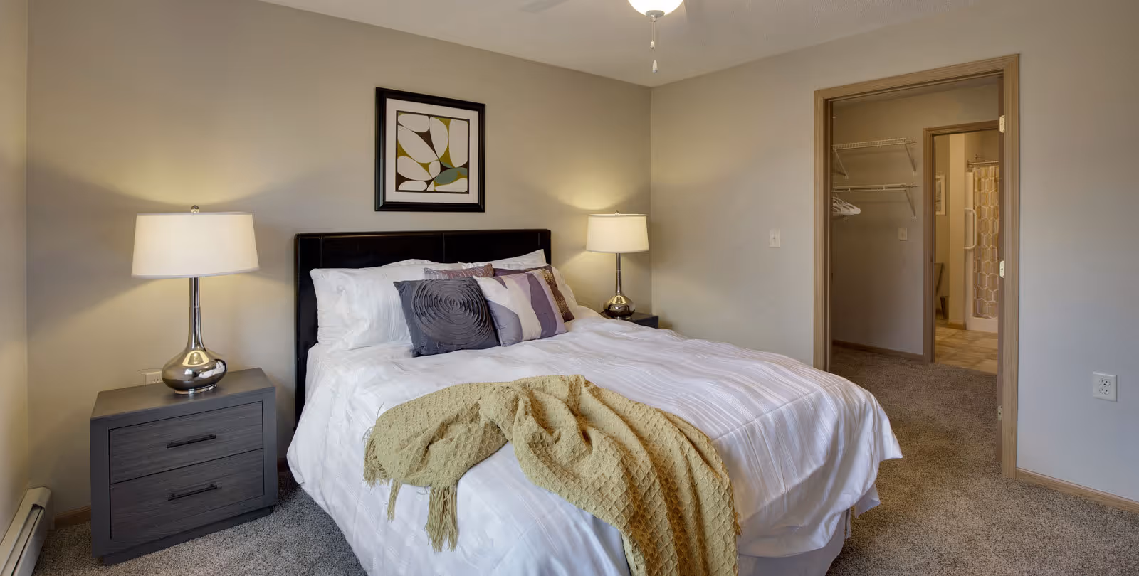 A neatly made bed with white linens and multiple decorative pillows in a bedroom. There is a yellow throw blanket draped over the foot of the bed. Two bedside tables with lamps flank the bed. A framed abstract artwork hangs on the wall above the headboard. An open doorway reveals a walk-in closet and a bathroom with a shower curtain.