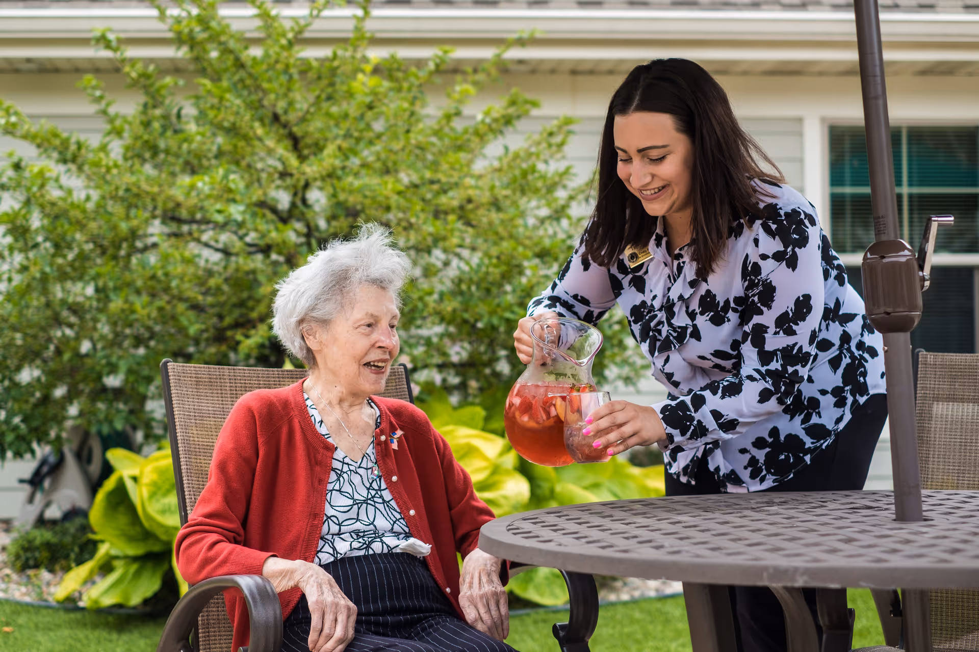 An elderly woman sitting outdoors at a patio table with a younger woman pouring a drink from a pitcher into a glass. The setting includes green plants and a building in the background.