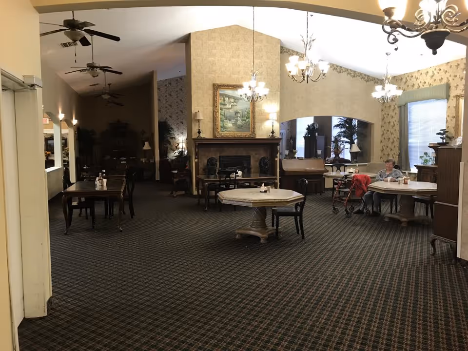 Interior view of a senior living facility common area with multiple tables and chairs, a fireplace with a framed painting above it, chandeliers hanging from the ceiling, ceiling fans, and a woman sitting at a table with a walker nearby. The room has patterned carpet and wallpaper with floral designs.