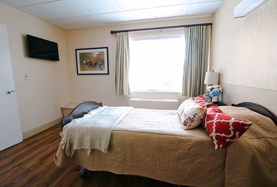 A neatly made single bed with beige bedding and decorative pillows in red and floral patterns in a well-lit room. The room has wooden flooring, a window with beige curtains, a wall-mounted flat-screen TV, a framed floral artwork, a bedside table with a lamp and a small flower arrangement.