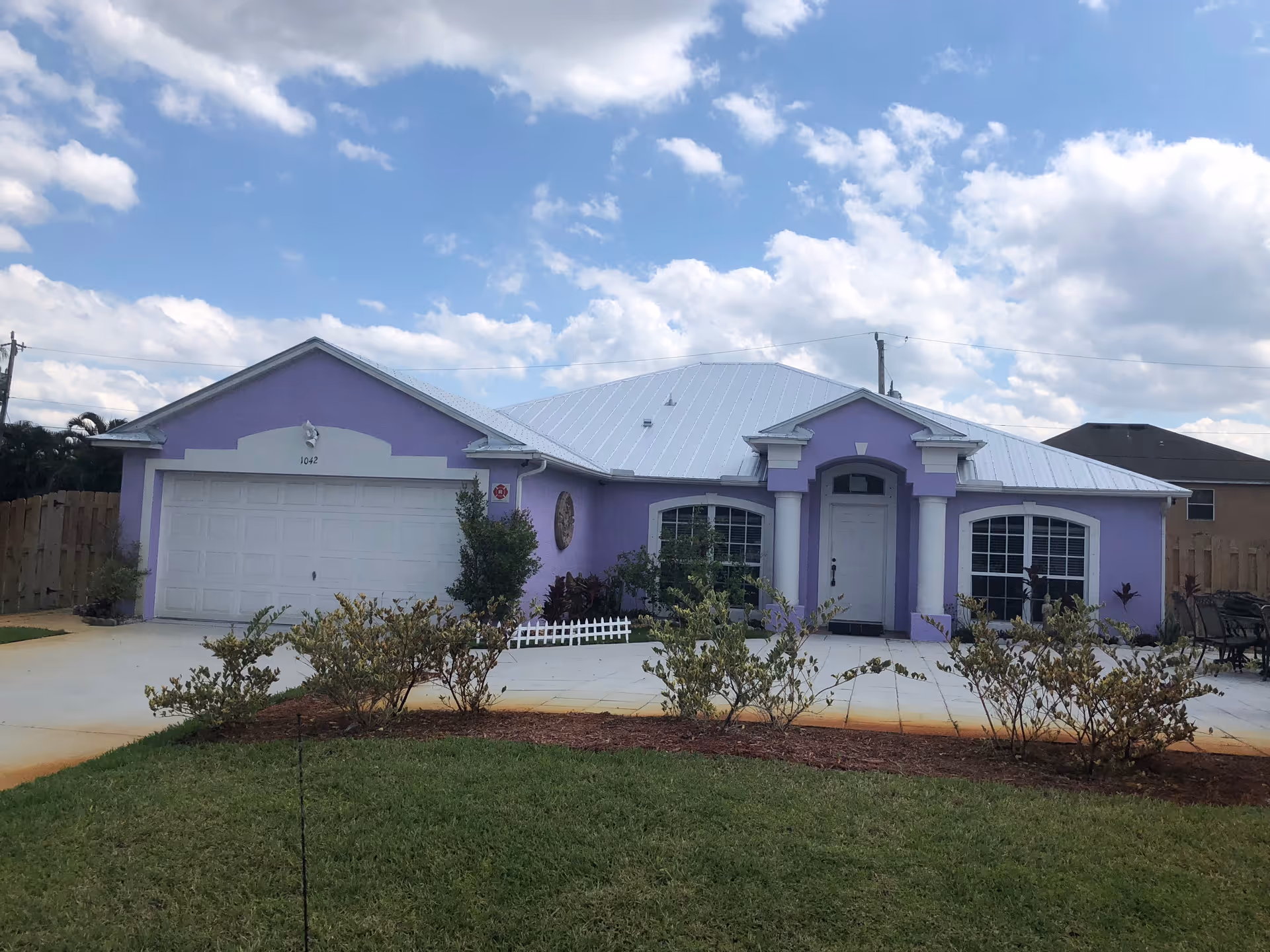 Front exterior view of a single-story purple house with a white metal roof, a two-car garage, and a driveway. There are shrubs and a small garden bed in front of the house, with a partly cloudy sky in the background.