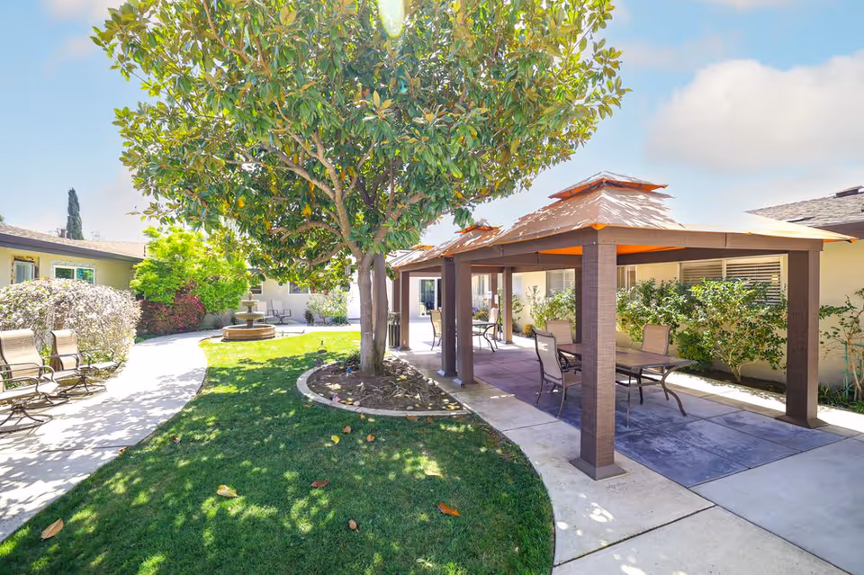 Outdoor courtyard area at St. John Kronstadt Health Care Center featuring a large tree, a covered seating area with tables and chairs, a curved walkway, green grass, and a water fountain in the background.
