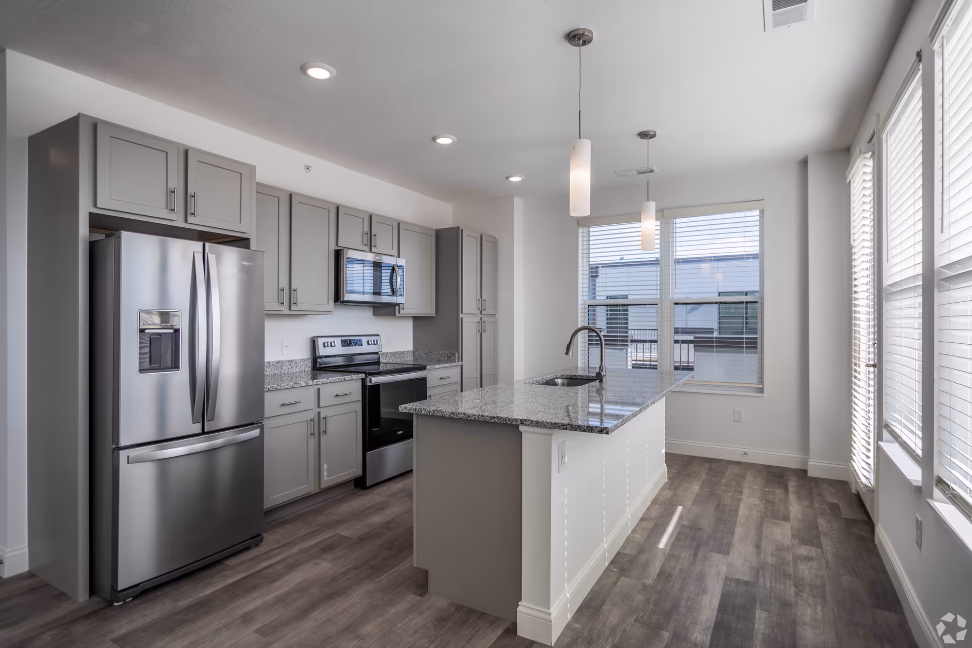 Modern kitchen with gray cabinets, stainless steel refrigerator, oven, microwave, and a large granite countertop island with a sink. The room has wood flooring and large windows with blinds allowing natural light to enter.