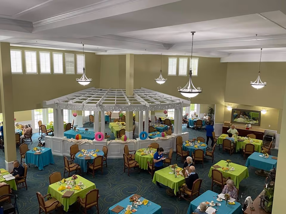 A spacious dining room in Cape Albeon Independent Living with multiple round tables covered in green and blue tablecloths. Several elderly residents are seated at the tables, some eating and others conversing. The room features a white pergola structure in the center decorated with string lights and colorful hanging ornaments. Large windows with white shutters allow natural light to fill the room, and elegant chandeliers hang from the ceiling.
