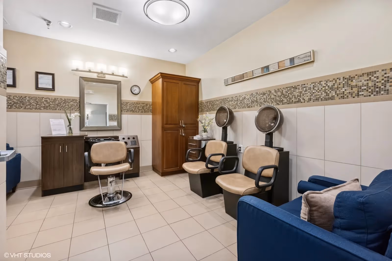 Interior view of a salon area with beige tiled floor and walls featuring a mosaic tile border. There are two beige salon chairs with black armrests in front of hair dryers, a single salon chair in front of a mirror and counter with cabinets, a wooden storage cabinet, and a blue cushioned chair with pillows on the right side.