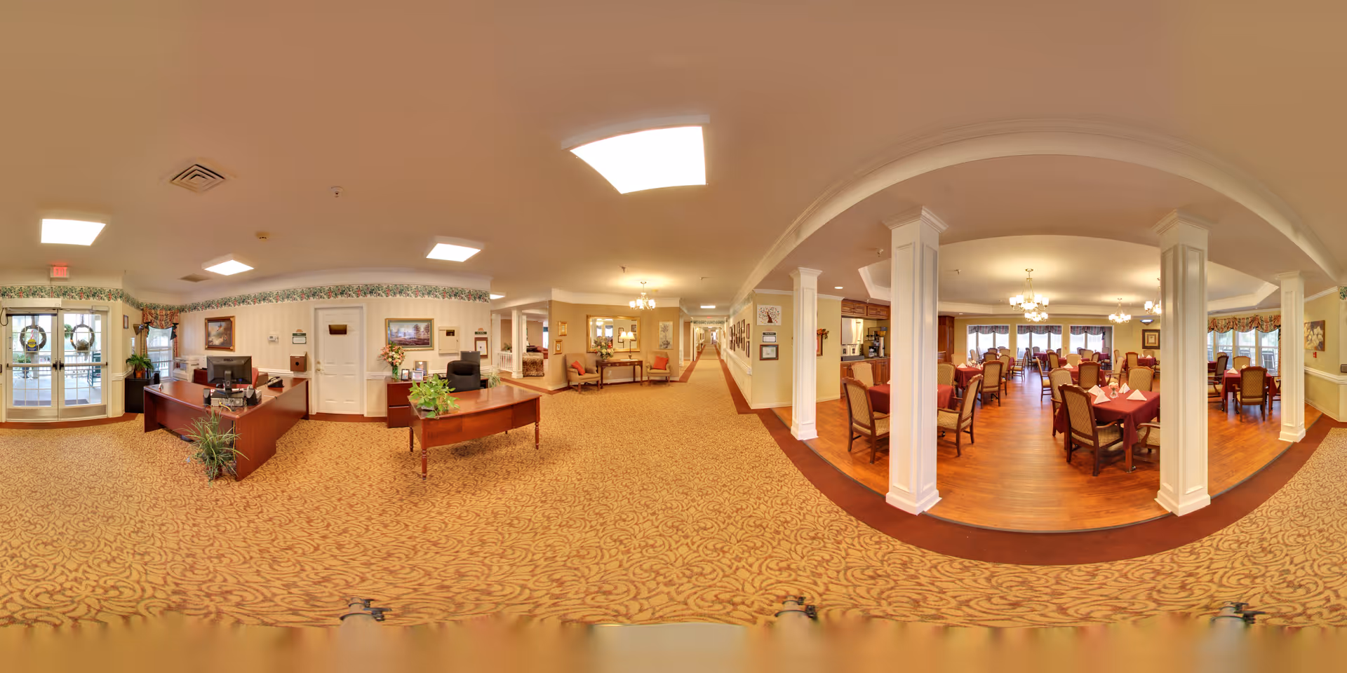 Panoramic view of the interior of Morningside of Auburn showing a reception area with desks and chairs on the left, a carpeted hallway in the center, and a dining room with multiple tables and chairs on the right. The dining room has wooden flooring and is decorated with chandeliers and window treatments. The reception area has plants and framed artwork on the walls.