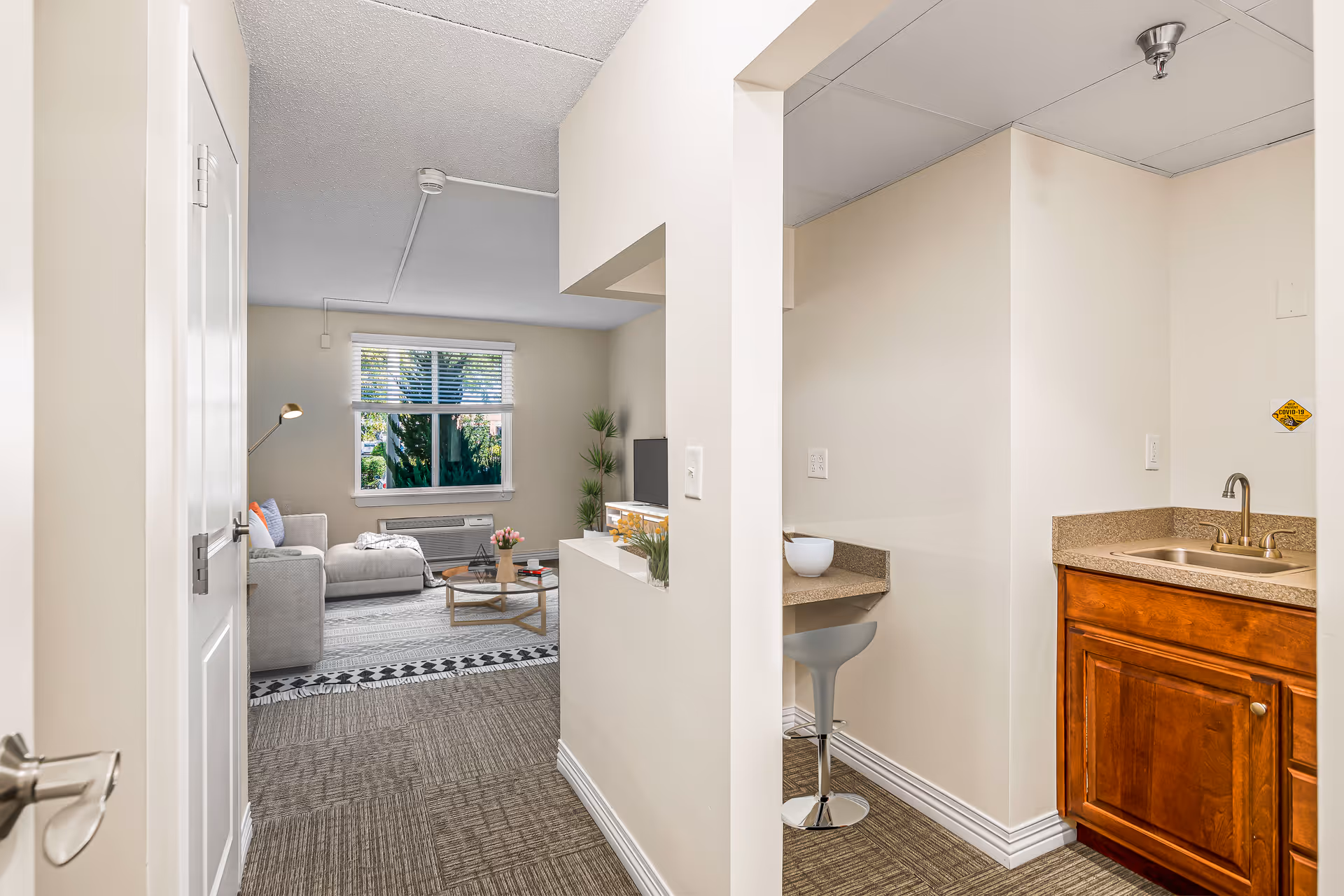 View from a hallway into a living area and a small kitchenette in a senior living facility. The living area features a gray sectional sofa, a coffee table with flowers, a floor lamp, a TV on a stand, and a window with blinds letting in natural light. The kitchenette has a countertop with a sink, wooden cabinets, and a small breakfast bar with a stool.