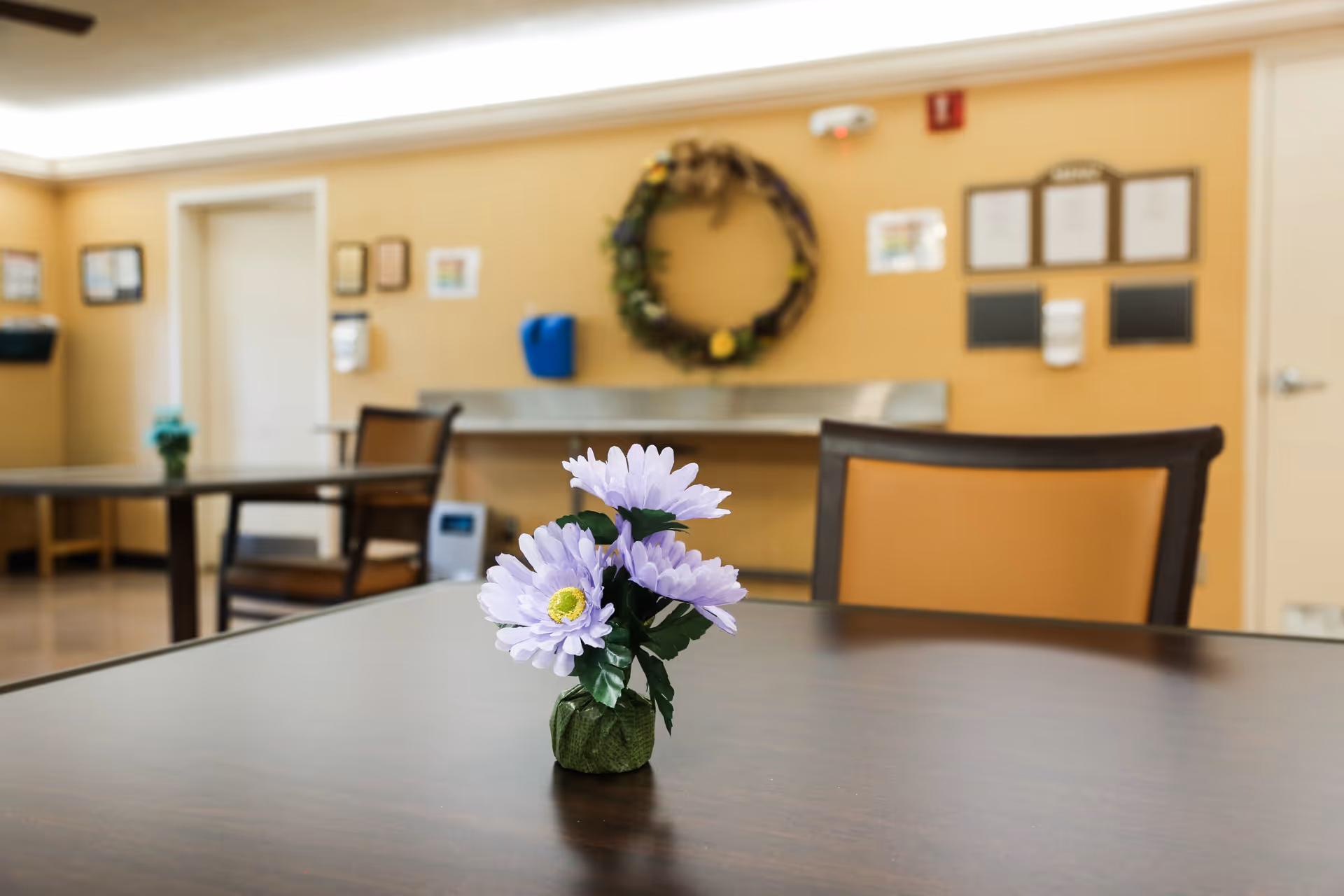 A small vase with purple flowers placed on a dark wooden table in a room with beige walls. In the background, there are chairs, a wreath hanging on the wall, and various wall-mounted items including a blue dispenser and framed documents.