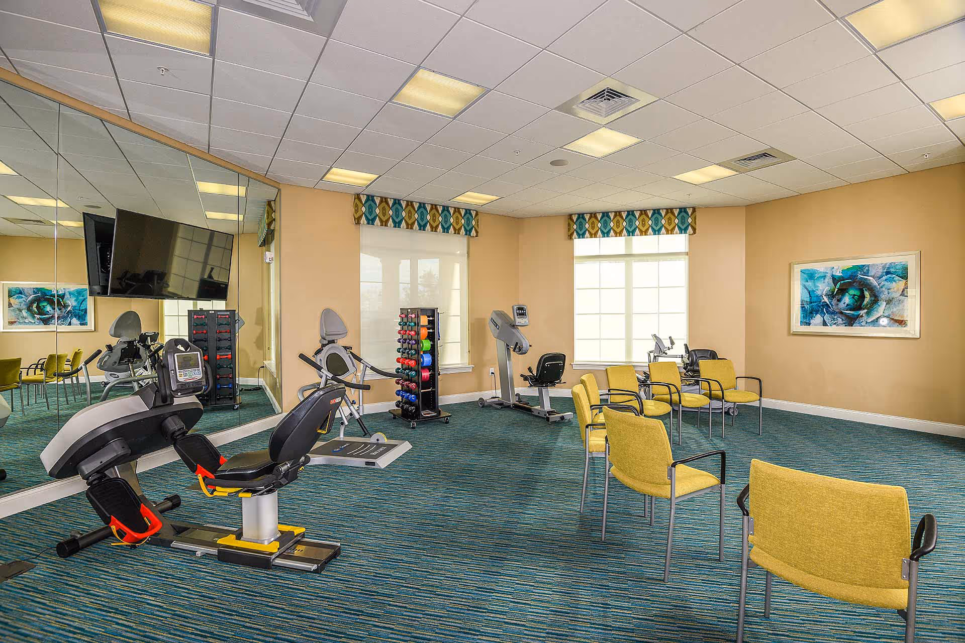 A fitness room with exercise equipment including a recumbent bike, a treadmill, and a rack of colorful dumbbells. The room has large windows with patterned valances, a wall-mounted TV, several yellow chairs arranged in a semi-circle, and a large mirror covering one wall.