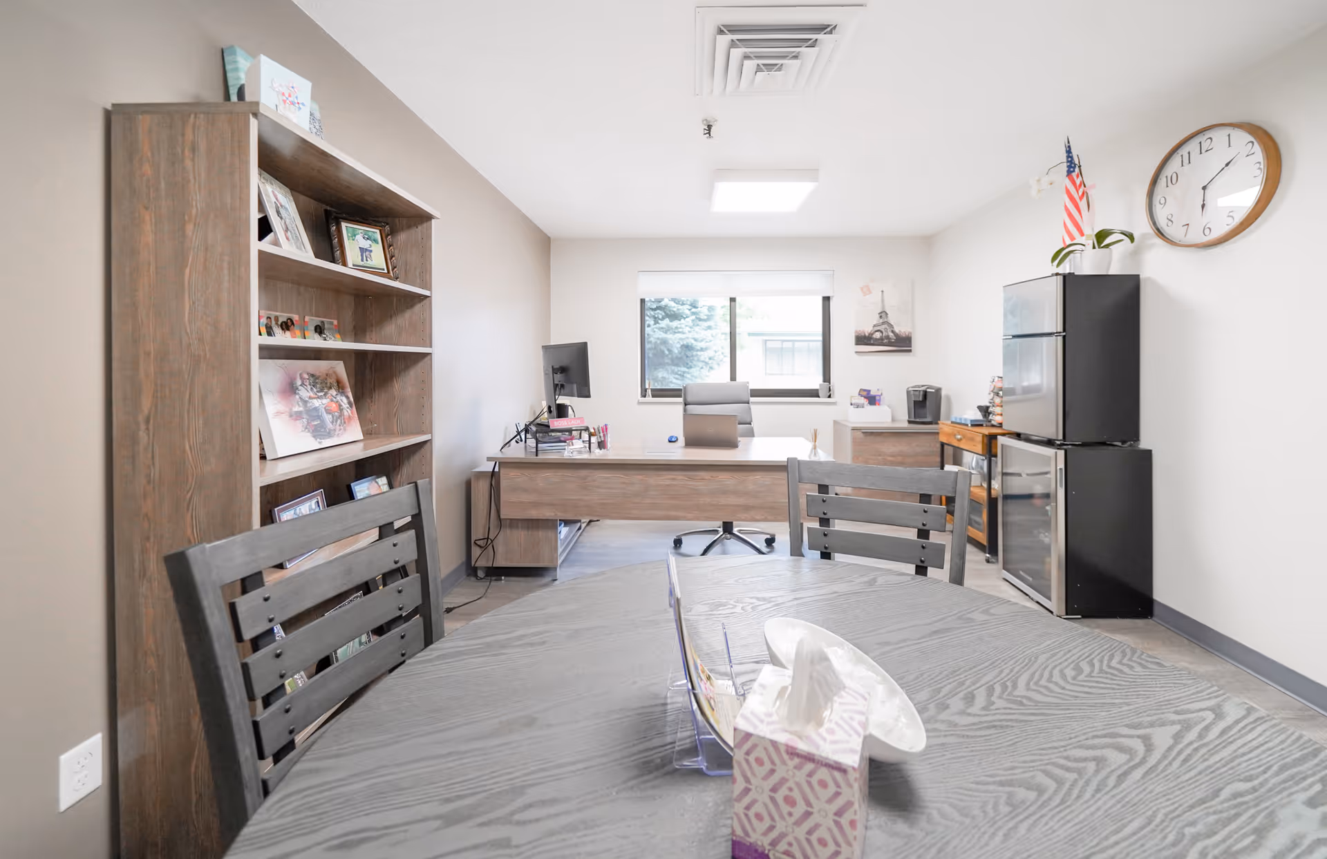 A bright office room with a wooden desk, office chair, computer monitor, and a bookshelf filled with framed photos and decorations. In the foreground, there is a round wooden table with chairs, a tissue box, and a clear organizer. On the right side, there is a black mini fridge with an American flag and a plant on top, a wall clock, and a window letting in natural light.