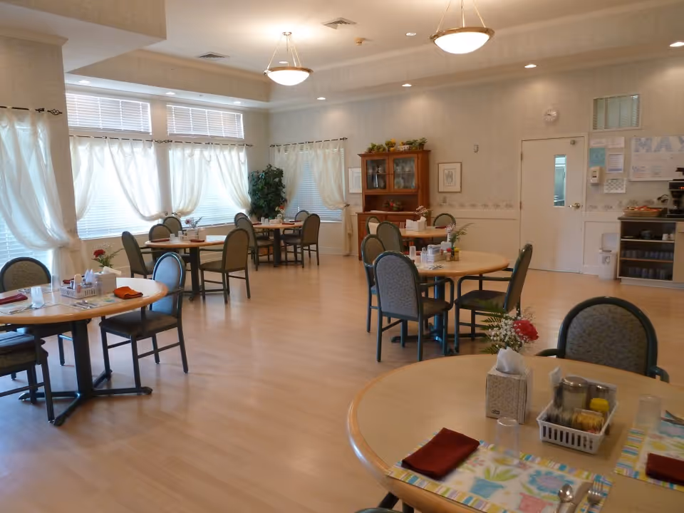 A bright dining room with several round tables and chairs arranged neatly. Each table has placemats, napkins, condiments, and small flower arrangements. Large windows with sheer white curtains allow natural light to fill the room. There is a wooden cabinet against the far wall and a bulletin board near a door. The floor is light wood, and ceiling lights provide additional illumination.