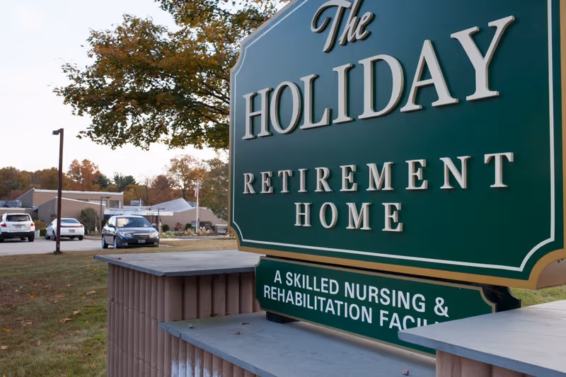 A large green sign for The Holiday Retirement Home, a skilled nursing and rehabilitation facility, with a background showing part of the building, parked cars, and trees with autumn foliage.