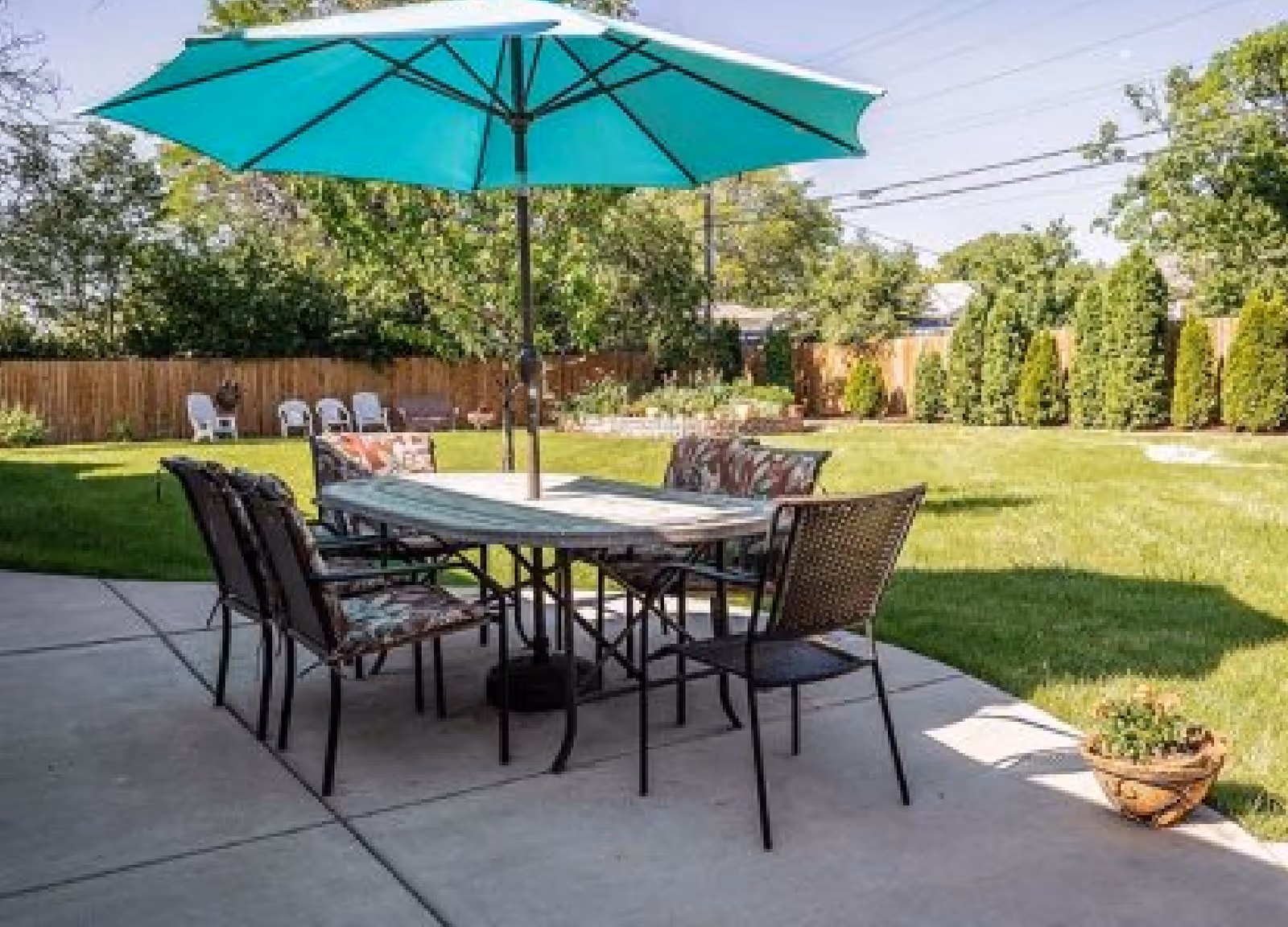 Patio dining table with chairs and a large turquoise umbrella on a concrete patio overlooking a grassy backyard with trees and a wooden fence.