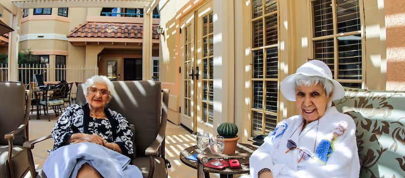 Two elderly women sitting comfortably on cushioned chairs in a sunny outdoor patio area with beige walls and large windows. One woman is wearing a black and white patterned top with a light blue blanket over her lap, and the other is wearing a white hat and a white sweater with colorful patterns. A small round table between them holds two glasses of water, a small potted cactus, and a pair of glasses.