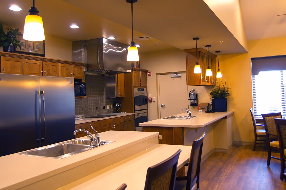 A modern kitchen area in Glen Carr House Memory Care featuring stainless steel appliances including a refrigerator and oven, wooden cabinets, two sinks with chrome faucets on beige countertops, pendant lights hanging from the ceiling, and a dining area with chairs near a window with blinds.