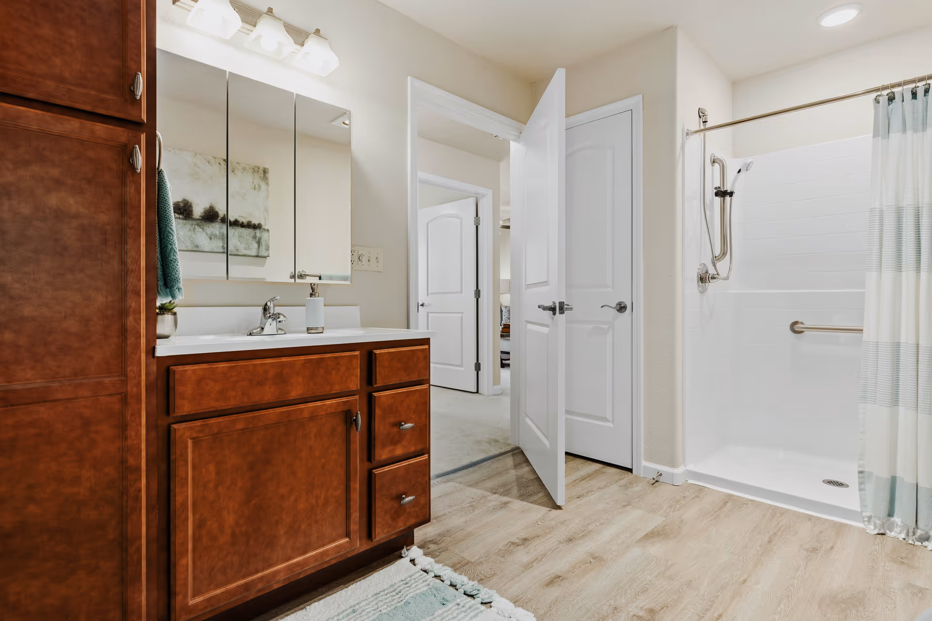 A clean and modern bathroom featuring a wooden vanity with a sink and mirror above it, a walk-in shower with grab bars and a striped shower curtain, light wood flooring, and an open door leading to another room.