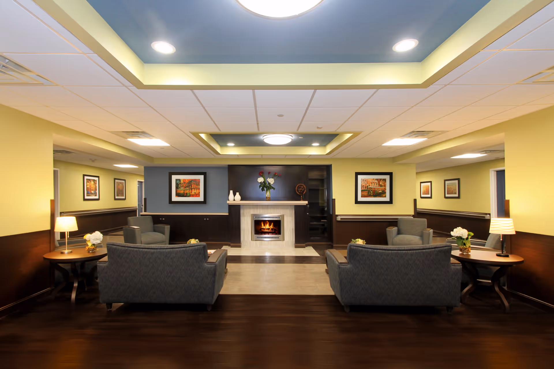 A cozy living room area in a senior living facility with two gray sofas facing a fireplace. The walls are painted yellow with dark wood paneling on the lower half. There are framed pictures on the walls, two side tables with lamps and flower arrangements, and a ceiling with recessed lighting and a central light fixture.