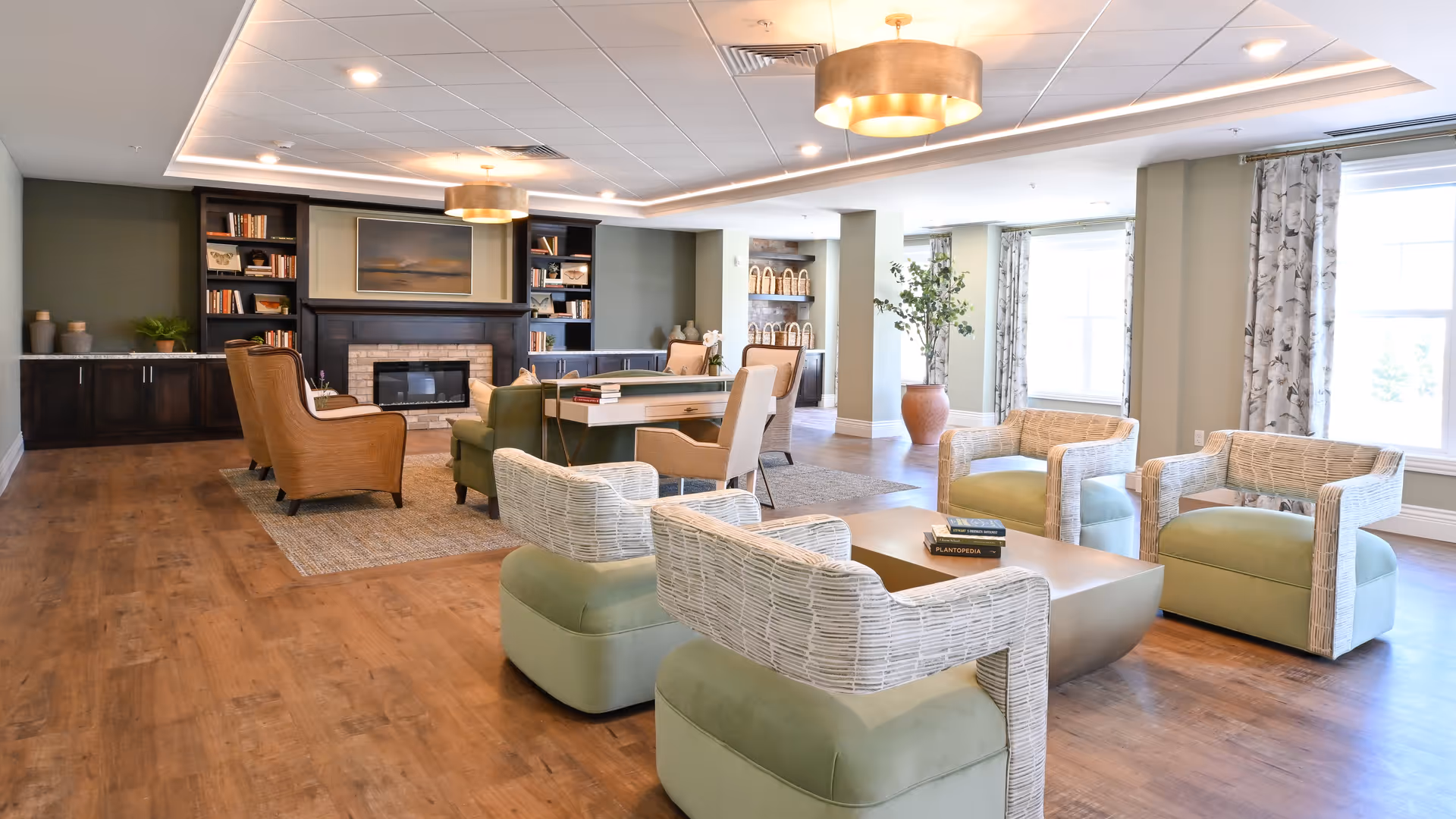 A spacious and well-lit living room area with wooden flooring, multiple comfortable chairs including green cushioned wicker chairs and tan armchairs, a central coffee table with books, built-in dark wood bookshelves flanking a fireplace, large windows with patterned curtains, and modern ceiling lights.