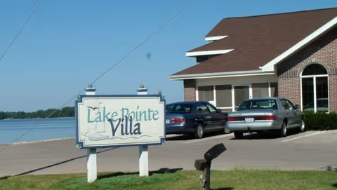 A sign reading "Lake Pointe Villa" stands on the lawn in front of a lakeside building with two parked cars.