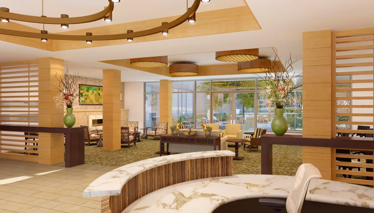 A spacious and well-lit senior living facility common area with modern decor. The room features a curved marble reception desk in the foreground, wooden pillars, and decorative vases with flowers. Comfortable seating areas with armchairs and small tables are arranged near large windows that let in natural light and offer a view of an outdoor patio with greenery. The ceiling has circular light fixtures, and the floor is a mix of tiled and carpeted areas.
