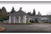 Exterior view of Providence Mother Joseph Care Center showing a single-story building with a covered entrance featuring white columns and a small cupola on the roof. The surrounding area includes a paved driveway, landscaped greenery, and trees in the background under an overcast sky.