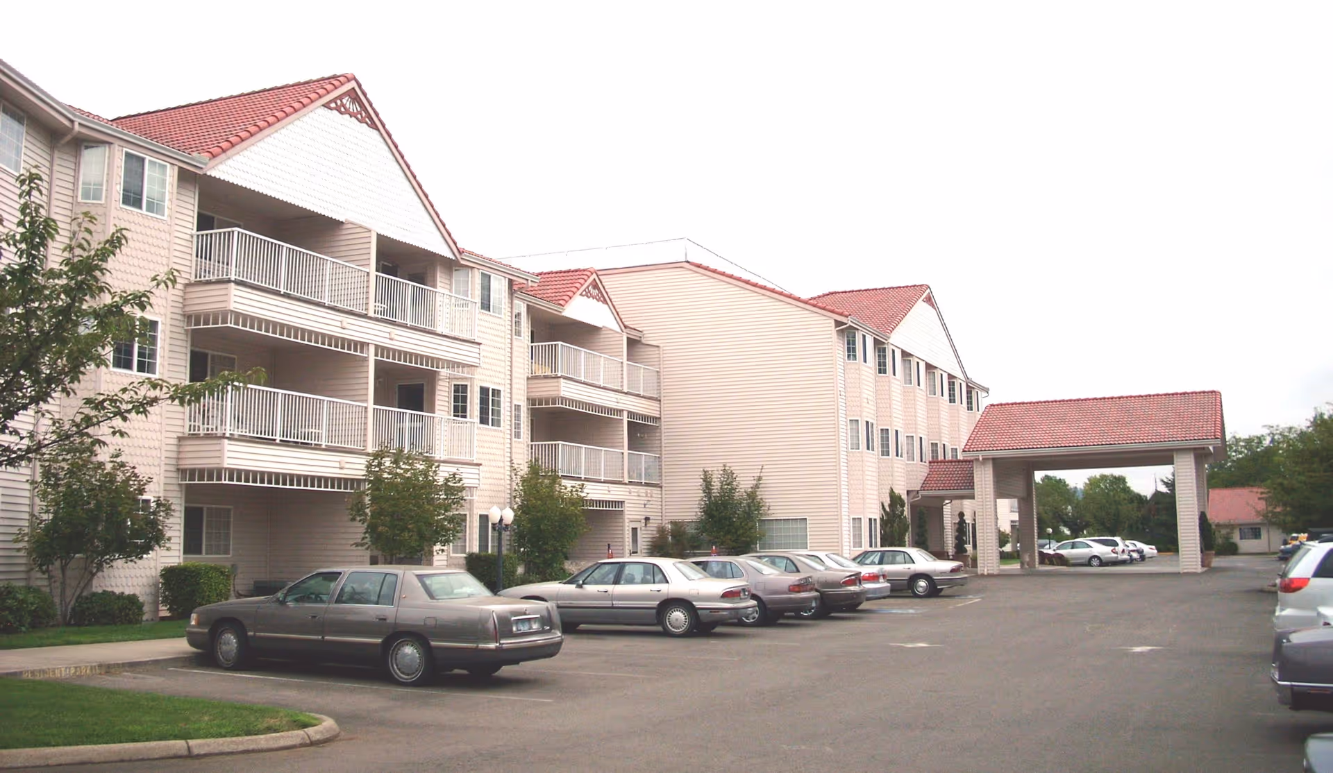Exterior view of Riverview Terrace senior living facility showing a three-story building with balconies, a covered entrance, and a parking lot with several parked cars.