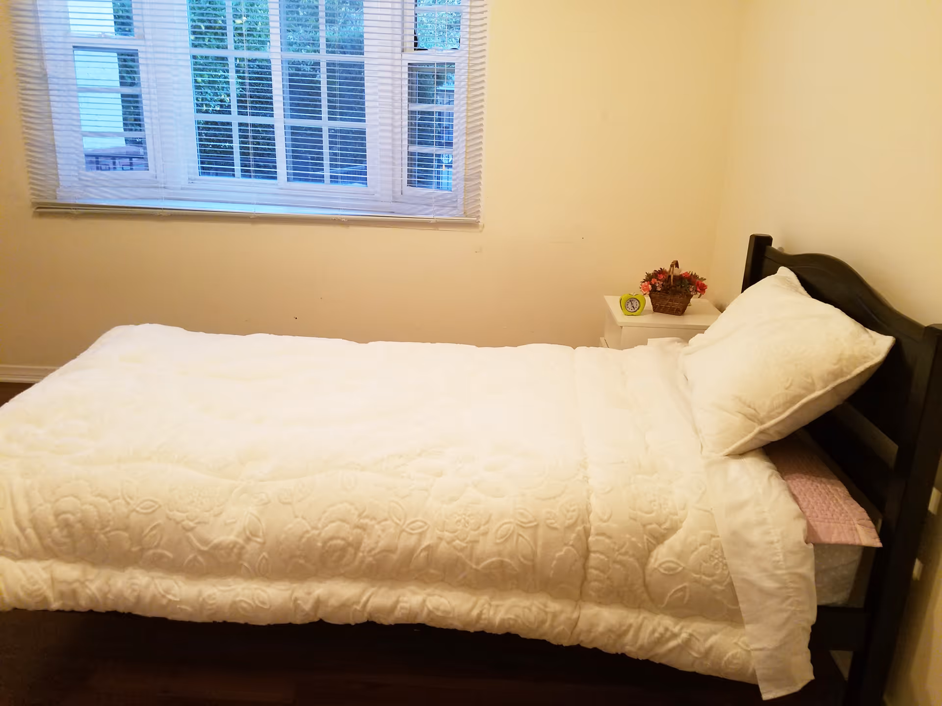 A small bedroom with a single bed dressed in white bedding, a pillow, nightstand with flowers, and a window with blinds.
