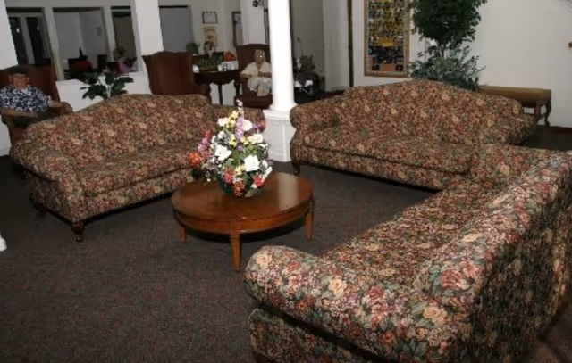 A common area in an assisted living facility featuring three floral-patterned sofas arranged around a wooden coffee table with a flower arrangement. In the background, two elderly individuals are seated in armchairs near white columns and a bulletin board.