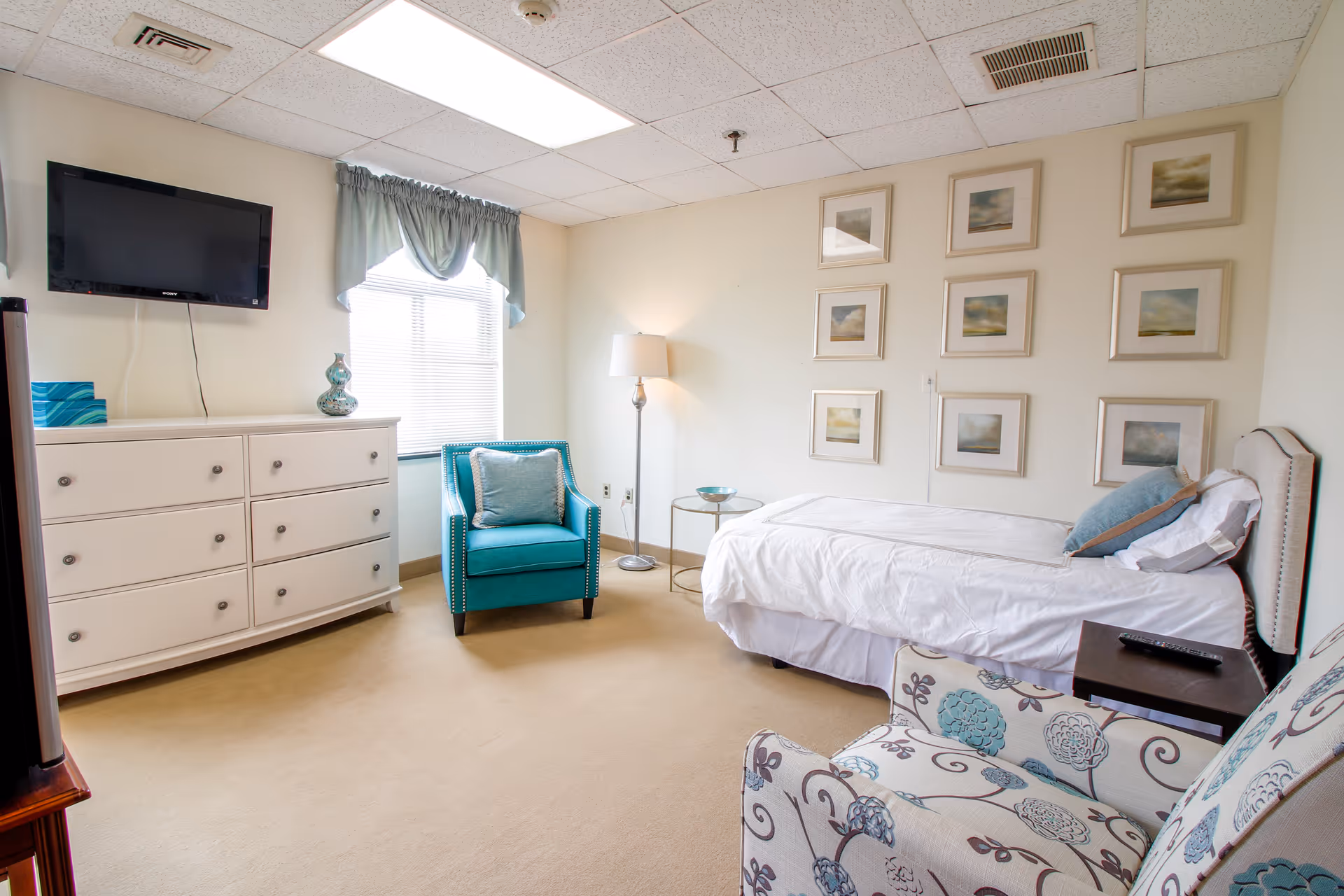 A bright and clean senior living bedroom featuring a single bed with white linens and blue pillows, a teal armchair with a matching pillow, a floral patterned armchair, a white dresser with decorative items, a wall-mounted flat-screen TV, a floor lamp, and nine framed pictures arranged in a grid on the wall.