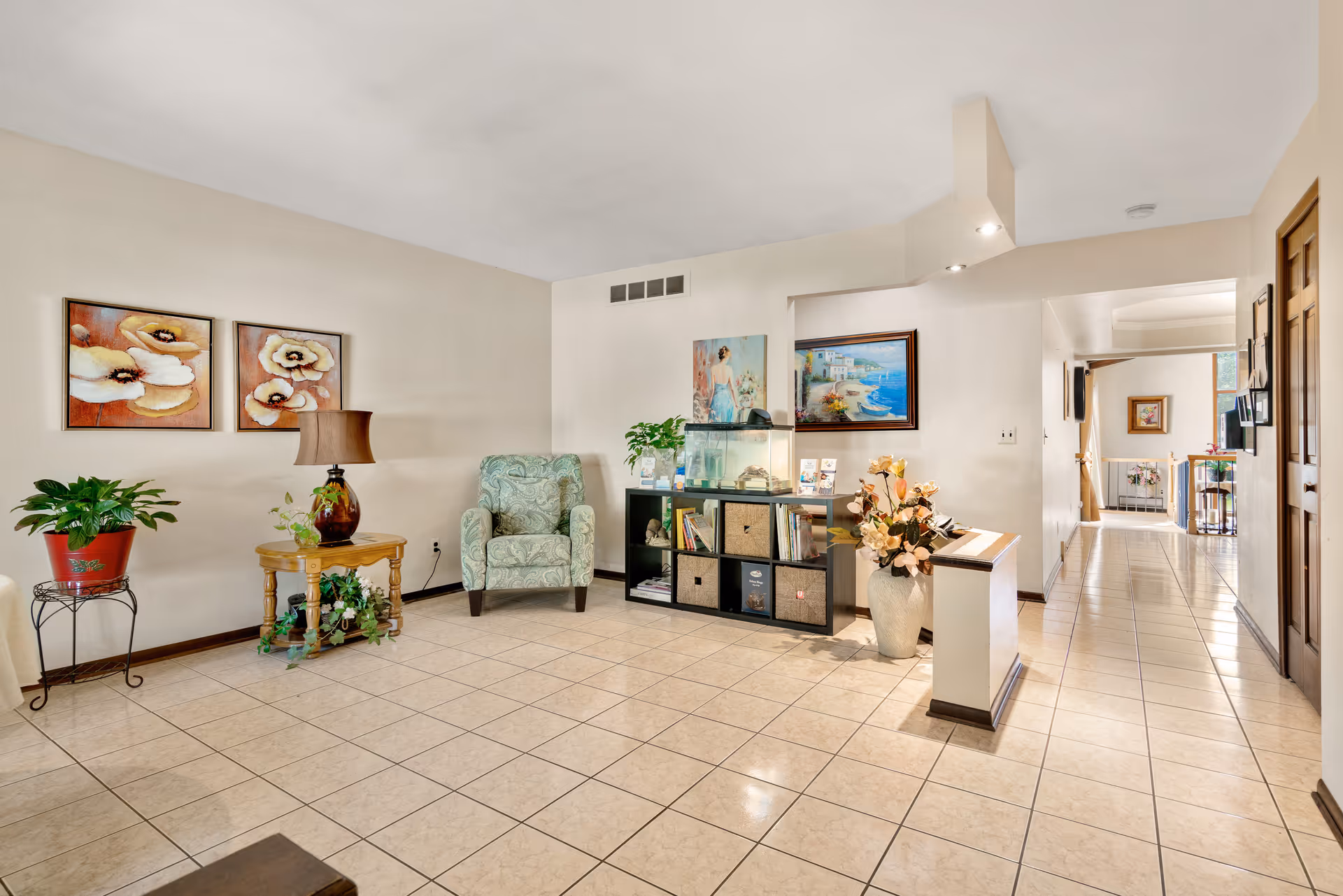 A bright and spacious interior room with tiled flooring, featuring a green patterned armchair, a wooden side table with a lamp and plants, a black shelving unit with baskets and books, an aquarium, and decorative paintings on the walls. The room leads into a hallway with more artwork and furniture visible in the distance.
