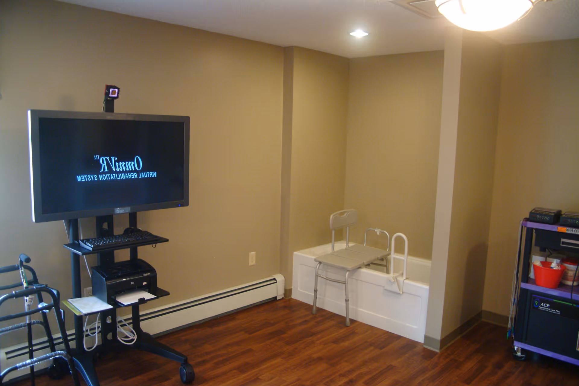 A room with beige walls and wood flooring featuring a mounted TV on a stand with a keyboard and printer below it. There is a walker on the left side and a bathtub with a shower chair inside it on the right side. A purple cart with various items is also visible on the far right.
