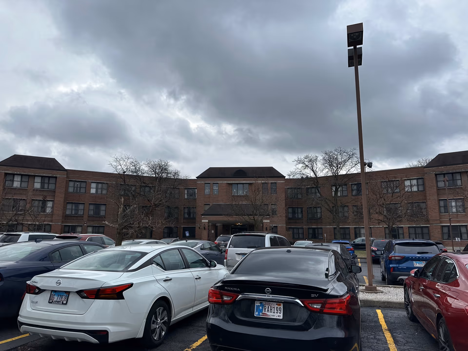 Exterior view of a three-story brick building with multiple windows, likely a nursing home, under a cloudy sky. Several cars are parked in the parking lot in front of the building, and there are leafless trees and a tall street lamp visible.