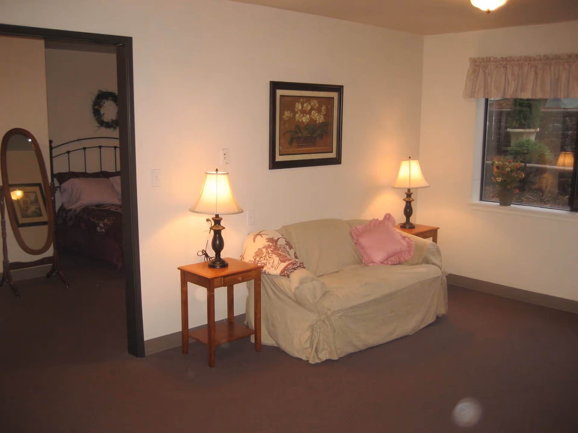 A cozy living room area with a beige loveseat covered with a slipcover and decorated with two pillows, one pink and one patterned. There are two wooden side tables on either side of the loveseat, each with a lit table lamp. A framed floral painting hangs on the wall above the loveseat. To the left, an open doorway leads to a bedroom with a bed, a standing mirror, and a wreath on the wall. A window on the right side shows some outdoor plants and a lamp reflection.