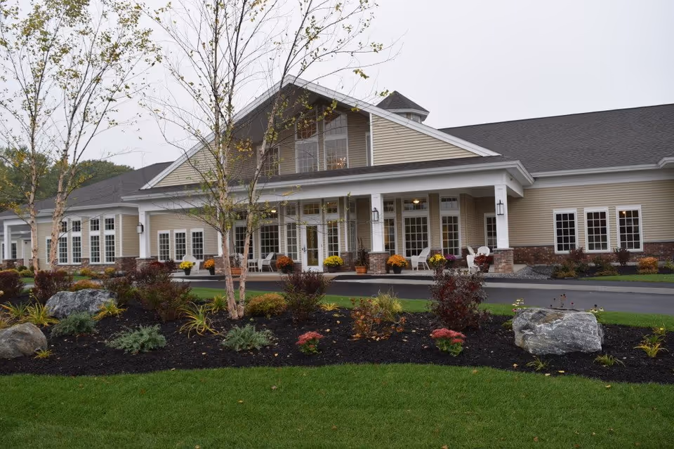 Exterior view of a single-story senior living facility building with beige siding, white trim, and a covered entrance porch. The landscaped front yard features green grass, small trees, shrubs, flowers, and large rocks. Several white chairs are placed on the porch.