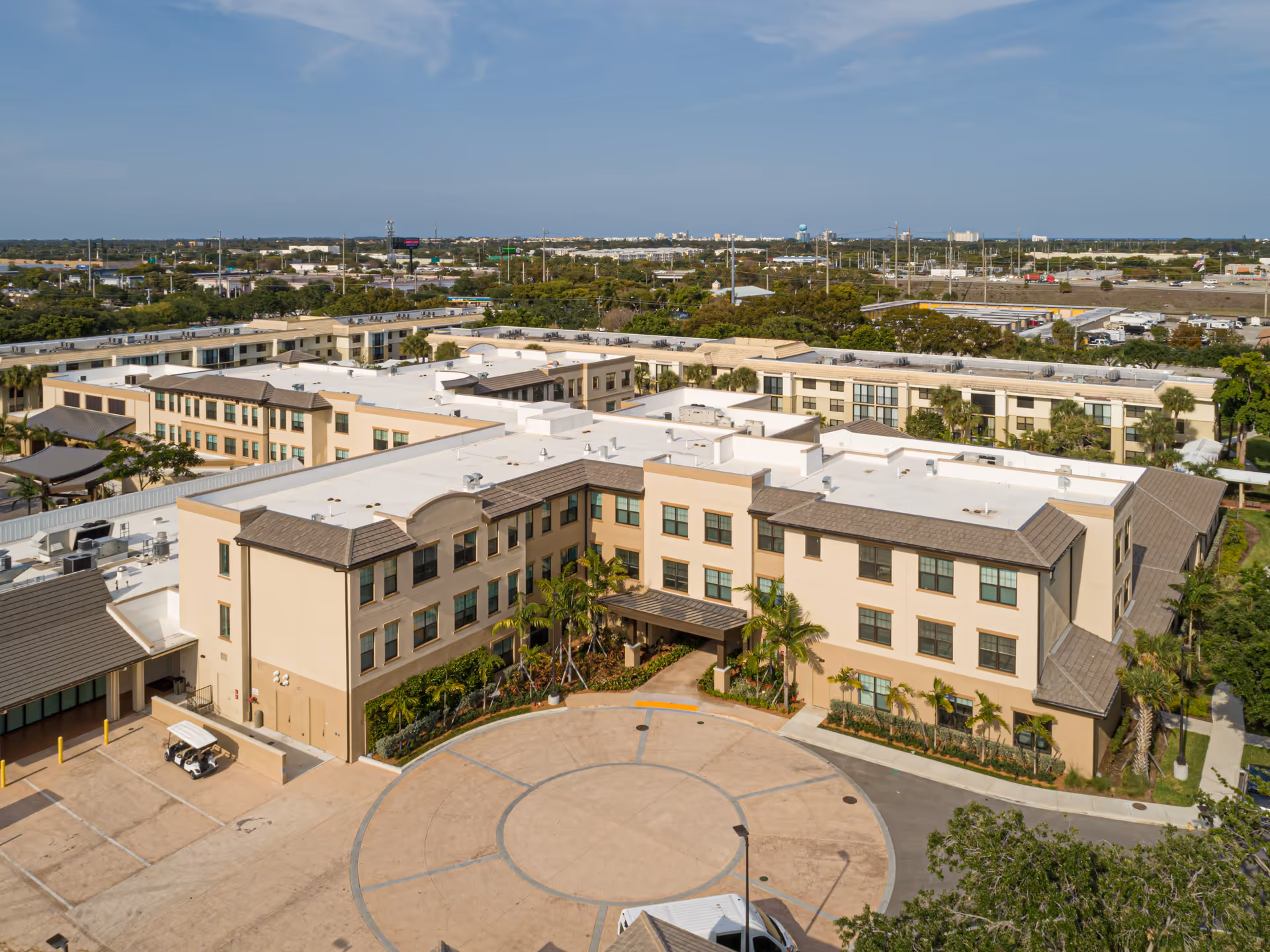 Aerial view of Cascades at Delray Beach senior living facility showing a large, multi-story beige building with a circular driveway entrance, surrounded by trees and greenery under a clear blue sky.