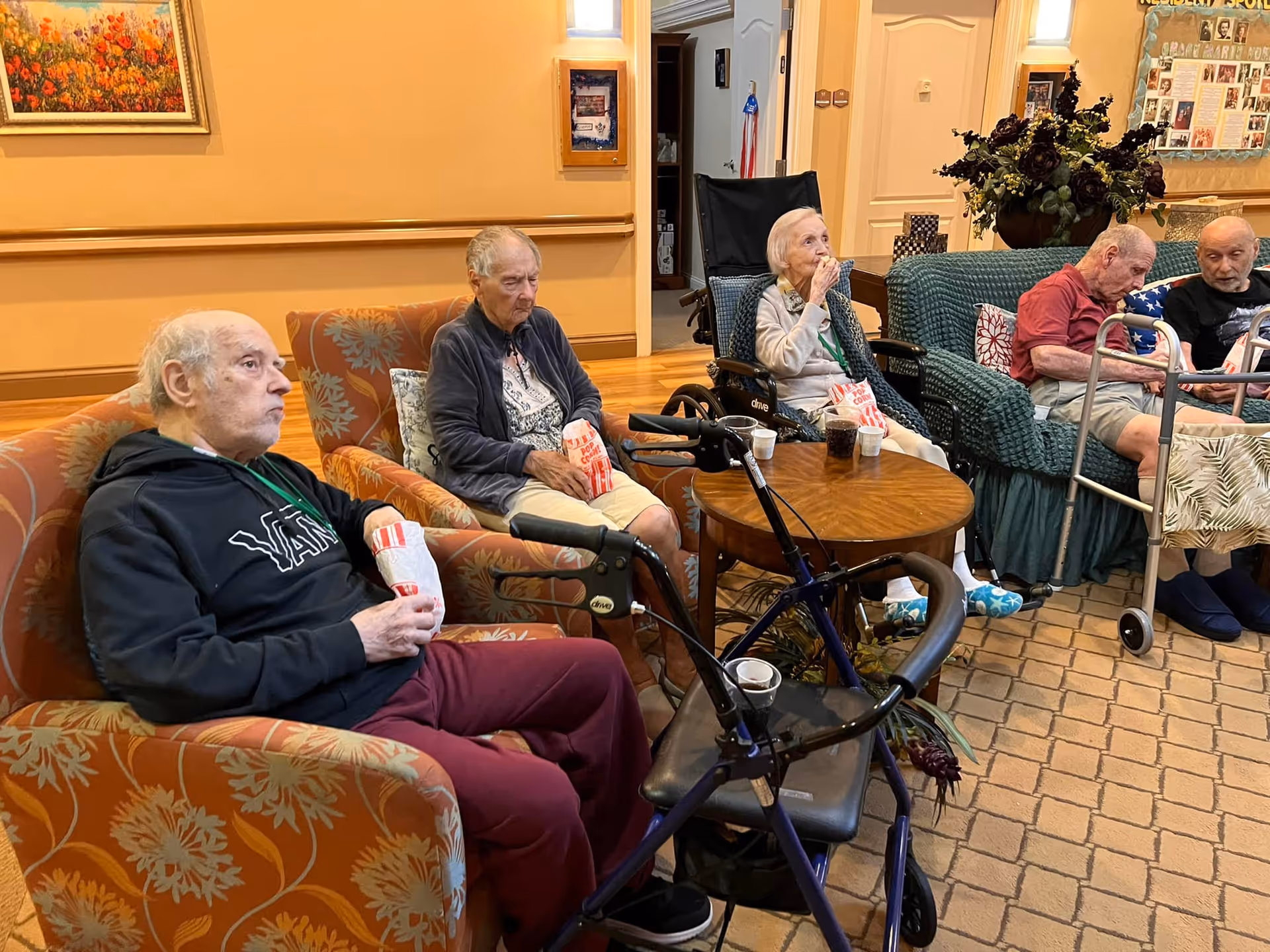 Five elderly individuals sitting in a cozy common area with armchairs and a sofa, some holding popcorn bags and drinks. Two walkers are visible in the foreground. The room has warm lighting, a framed painting on the wall, and a bulletin board in the background.