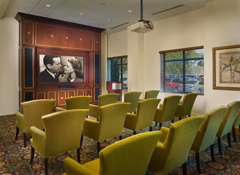Small screening room with rows of green upholstered chairs facing a wall-mounted black-and-white movie on a wood-paneled wall and a ceiling projector.