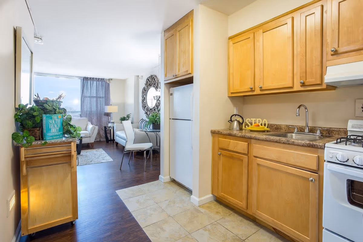 View of a small kitchen with wooden cabinets, a white refrigerator, a stove, and a sink. The kitchen opens into a living area with a dining table and chairs, a sofa, a large decorative mirror on the wall, and a window with sheer curtains letting in natural light.