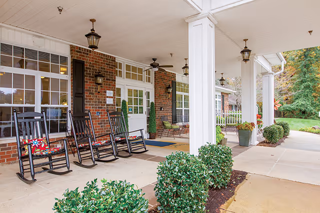 Covered front entrance porch of a senior living facility with rocking chairs, white columns, and a brick facade.