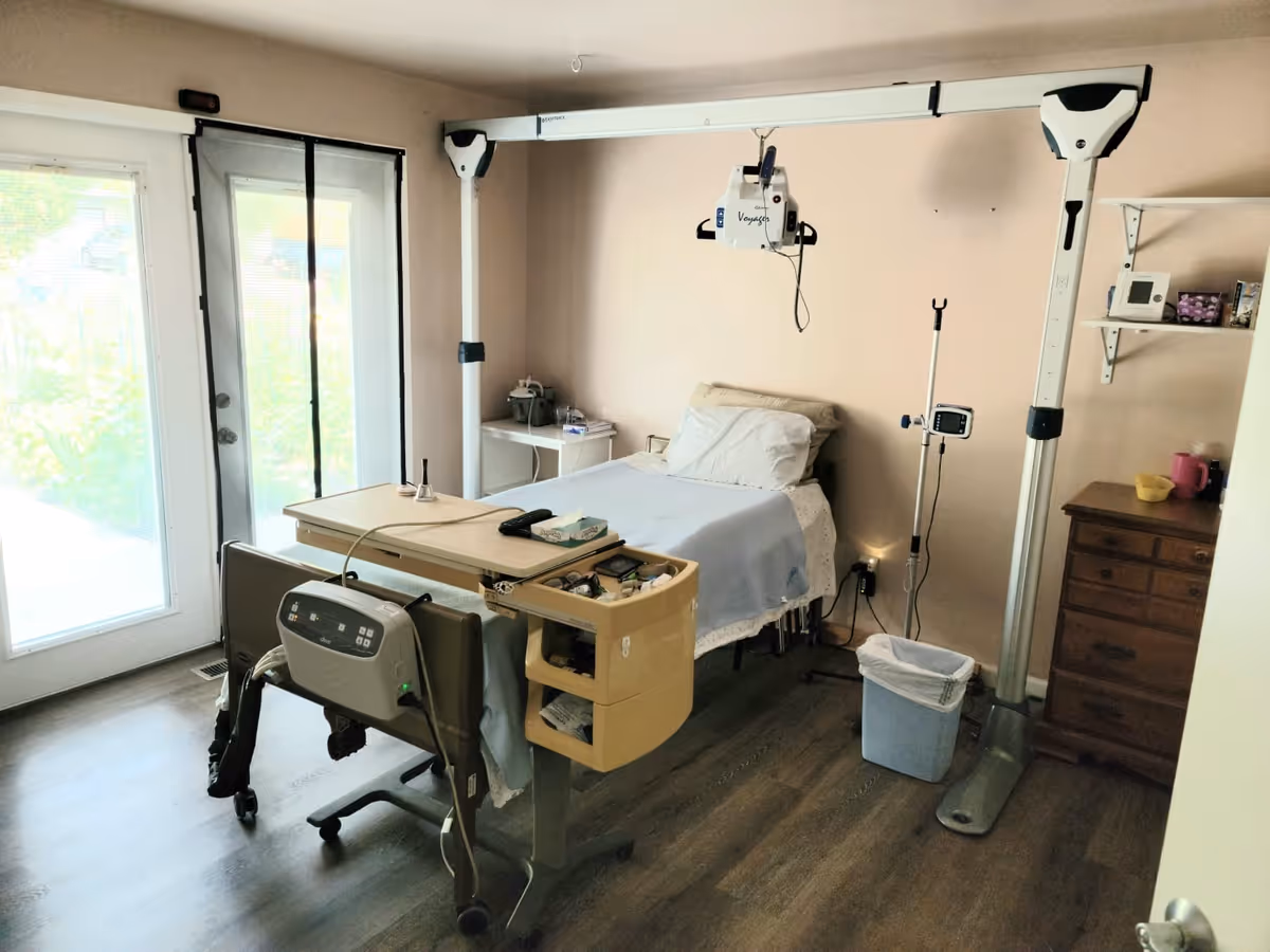 A private bedroom with an adjustable hospital bed, ceiling-mounted patient lift, IV pole, dresser and sliding glass door to the outside.