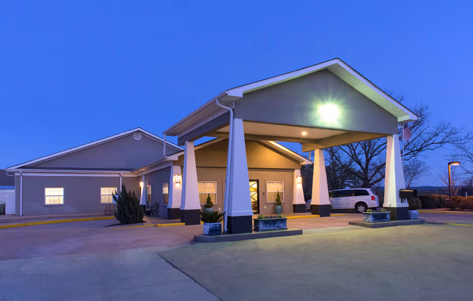 Exterior view of Quapaw Nursing & Rehab Center at dusk, showing the building entrance with a covered drop-off area supported by white pillars, illuminated by outdoor lights. A white vehicle is parked near the entrance, and trees and bushes are visible in the background.