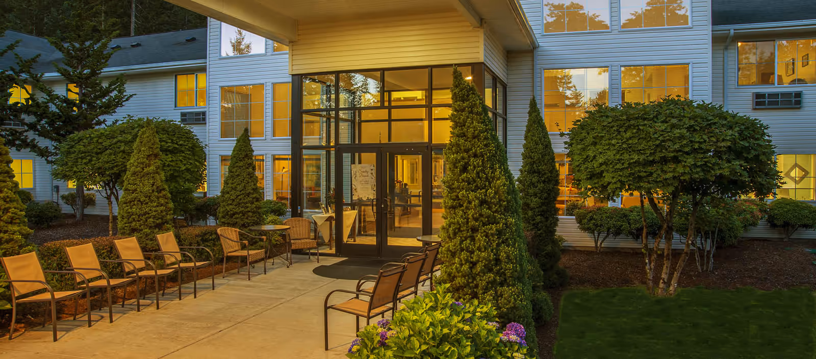 Evening view of the entrance to Maple Glen Assisted Living facility with a pathway lined by chairs and surrounded by well-maintained shrubs and trees. The building has large windows illuminated from inside.