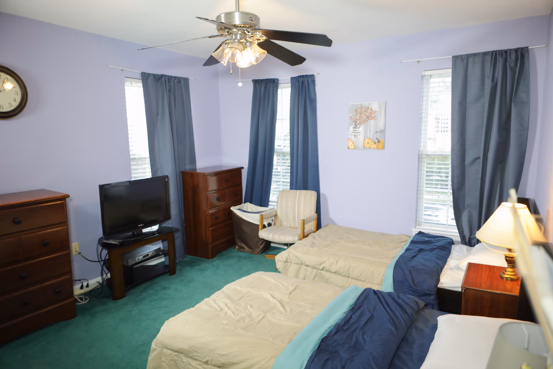 A light purple-walled bedroom with two twin beds, wooden dressers, a TV, an armchair, blue curtains, and a ceiling fan.
