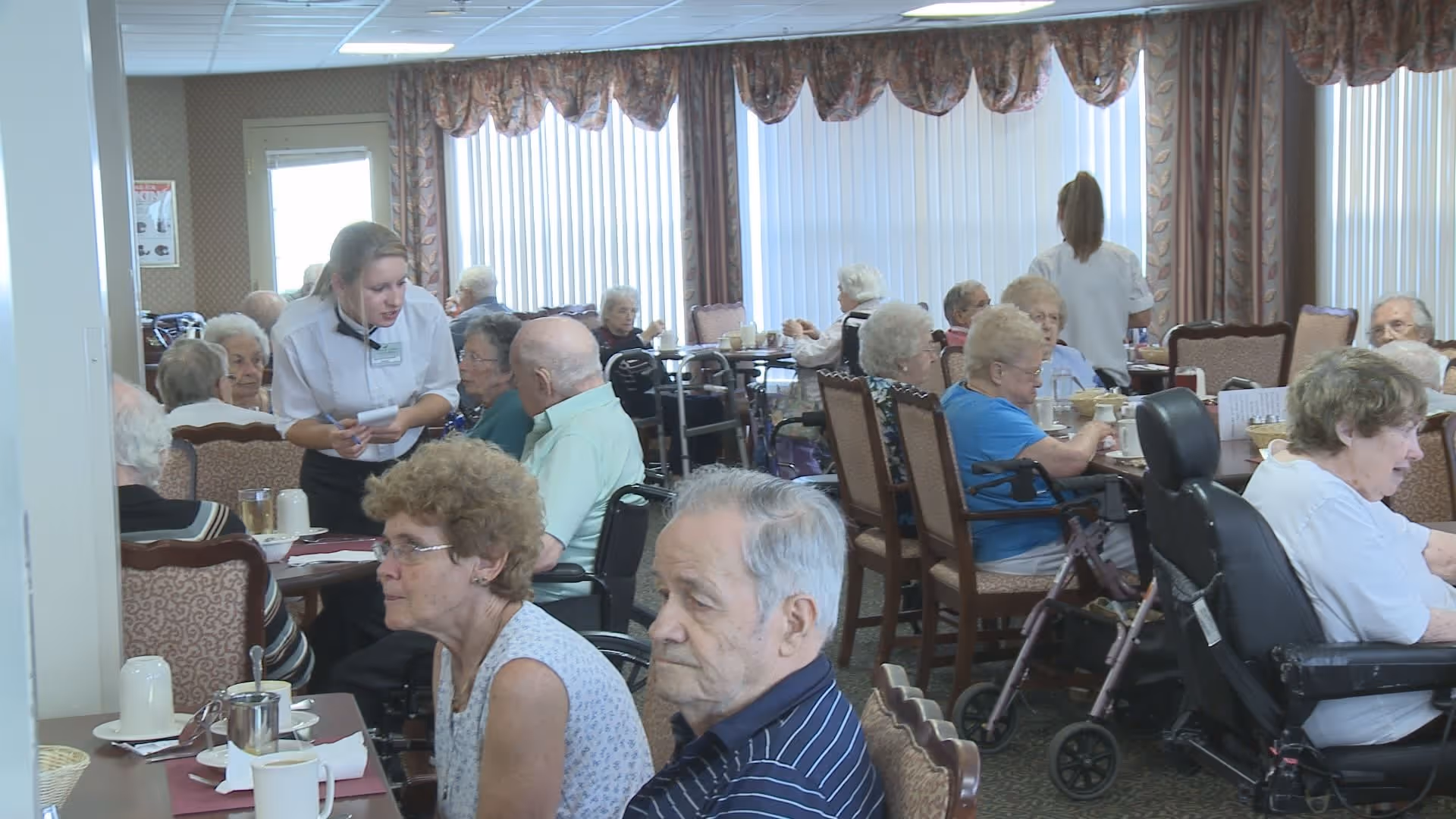 A dining room in a senior living facility with elderly residents seated at tables, some in wheelchairs, eating and socializing. Two staff members are attending to the residents. The room has large windows with vertical blinds and patterned curtains.