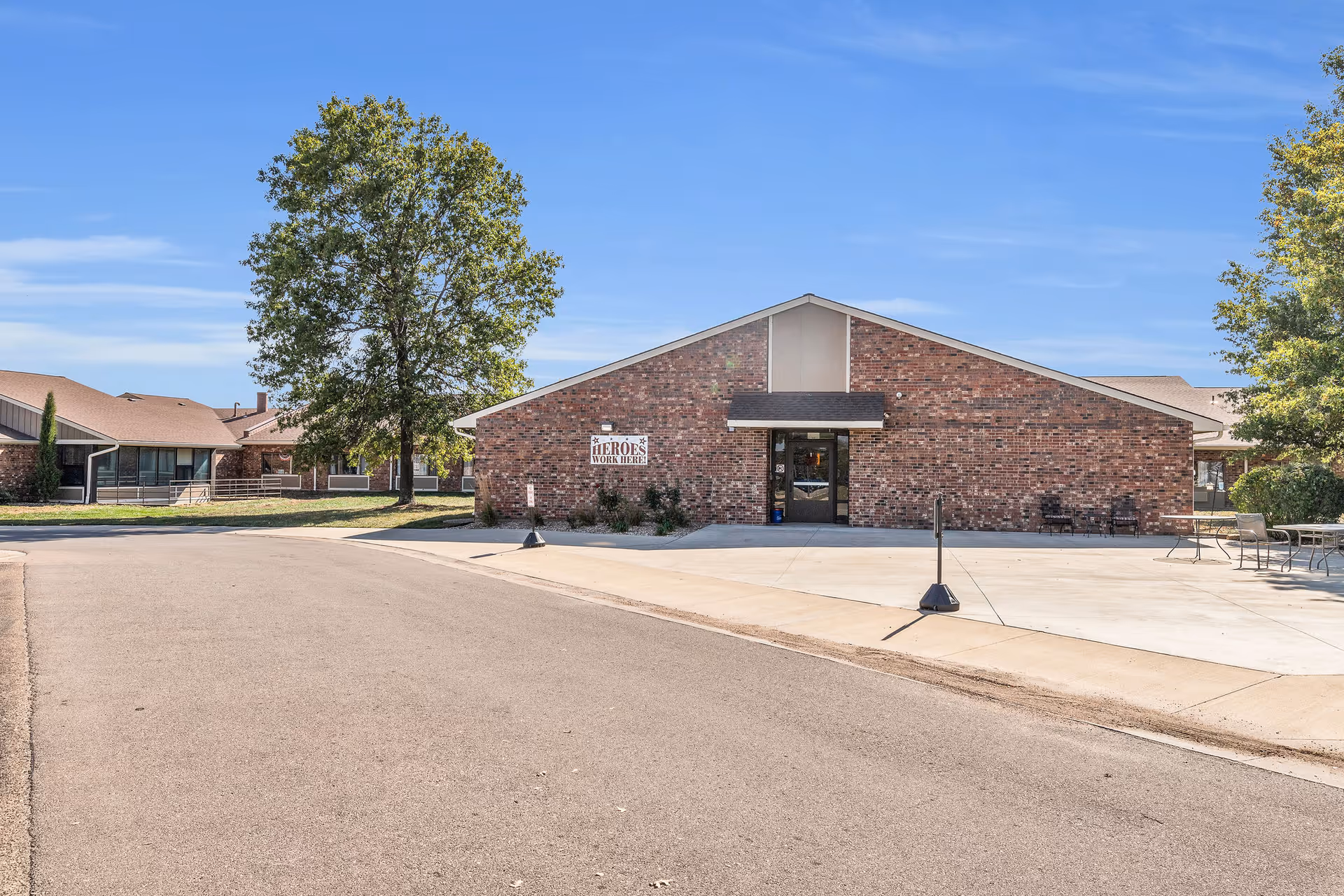 Front exterior of a single-story brick senior living facility with a paved driveway, patio seating, and trees under a blue sky.