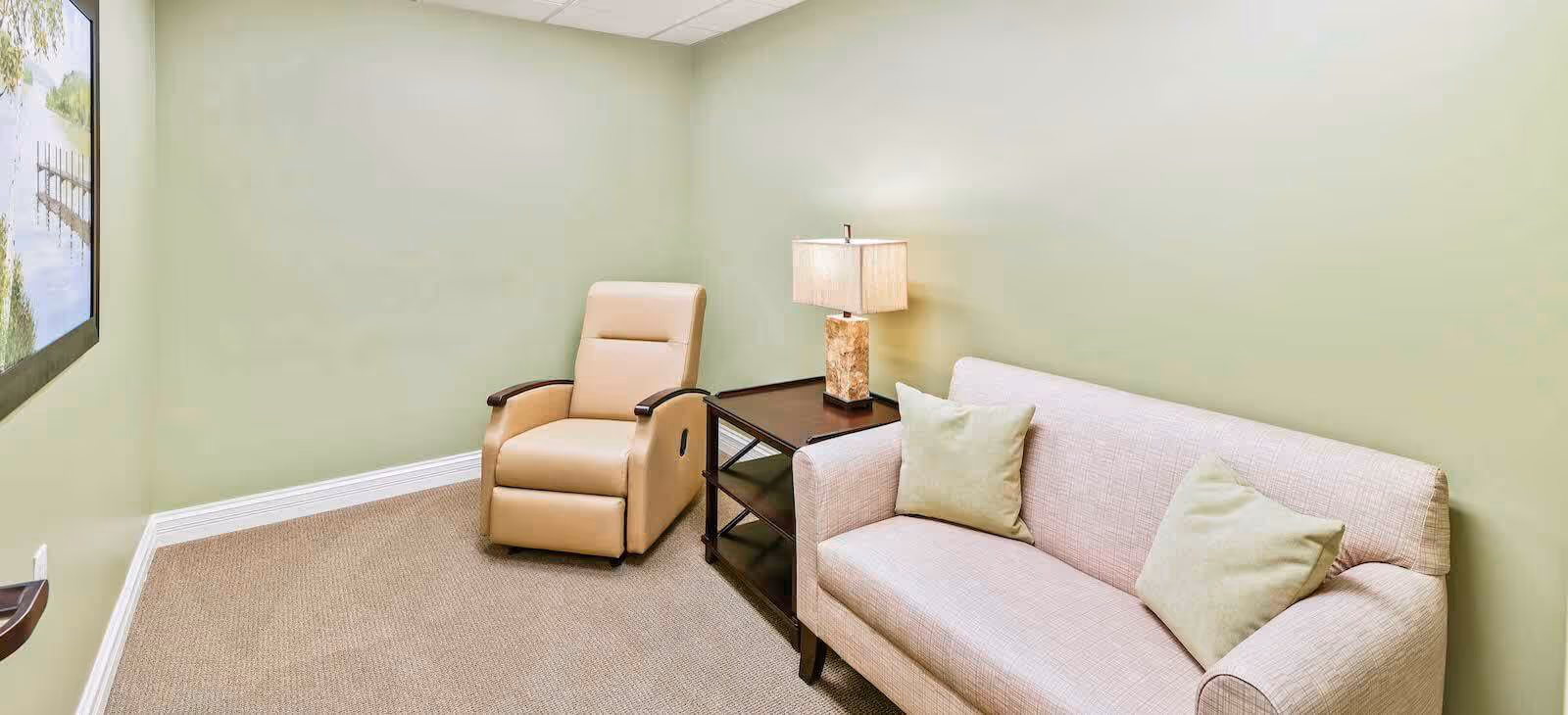 Small sitting room with a beige recliner, light sofa with pillows, a side table and lamp against pale green walls.