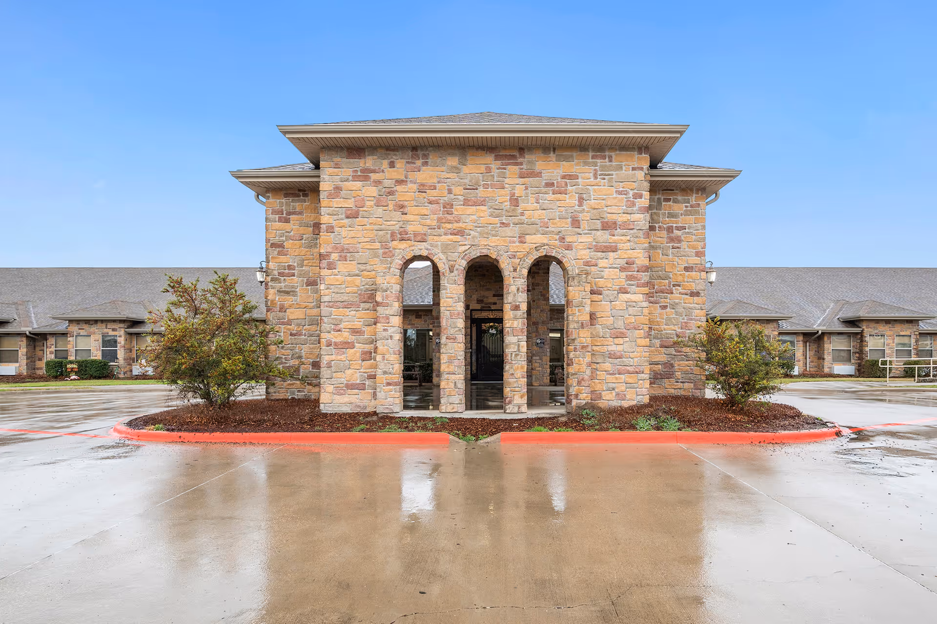 Front exterior view of a nursing and rehabilitation facility with a stone facade featuring three arched entryways, flanked by small landscaped areas with bushes, and a wet concrete driveway in front under a clear blue sky.