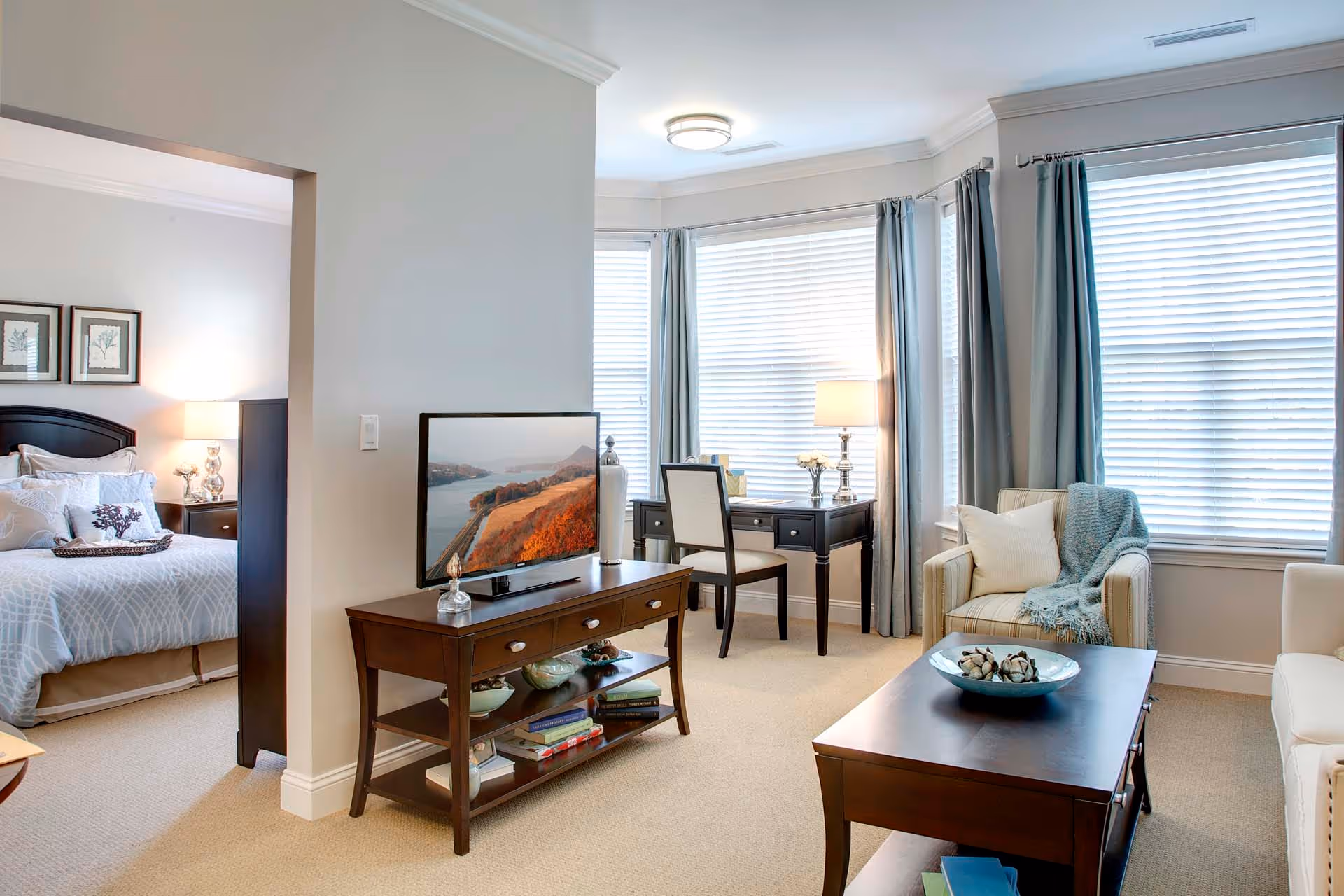 A bright and cozy living room area with a TV on a wooden stand, a coffee table with a decorative bowl, an armchair with a throw blanket and pillow, and a desk with a chair and lamp near large windows with blinds and curtains. A bedroom with a bed, nightstand, and lamp is visible through an open doorway.