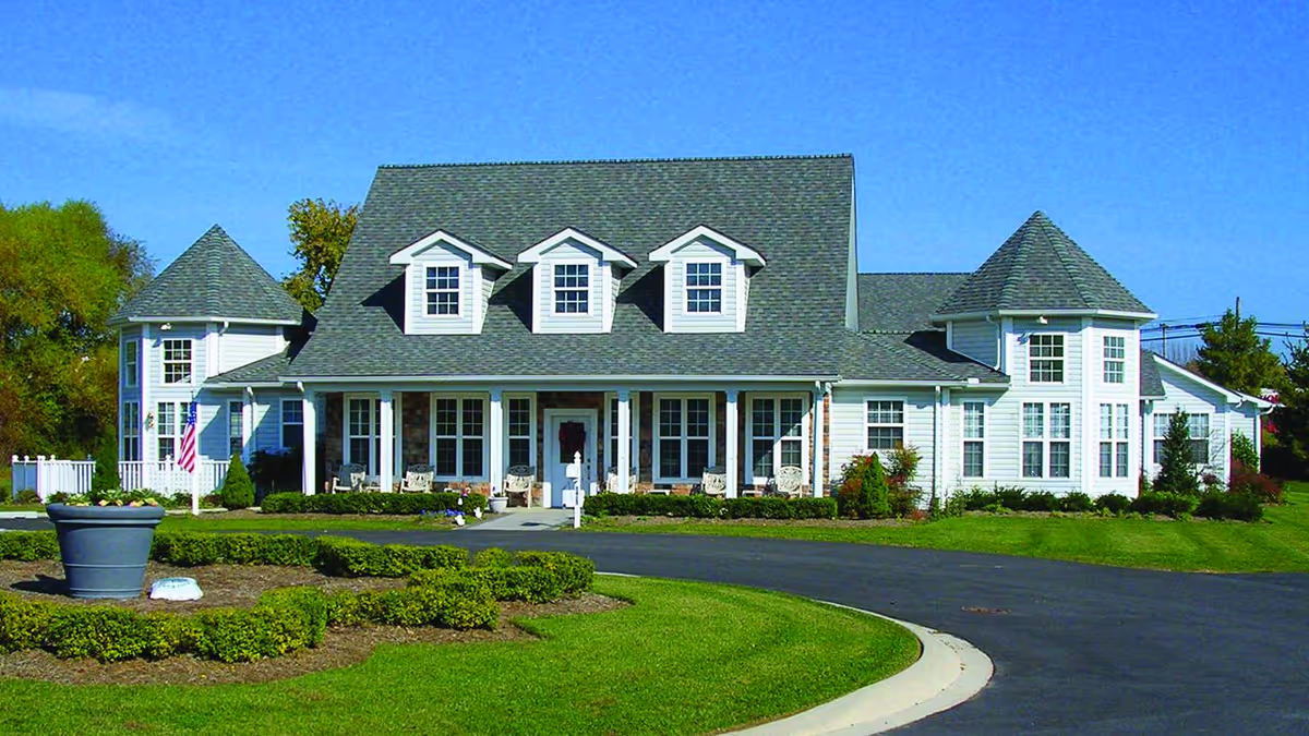Front exterior view of a large, white senior living facility building with a gray shingled roof, multiple windows, and a covered porch with chairs. The building is surrounded by well-maintained green lawns, shrubs, and a circular driveway.