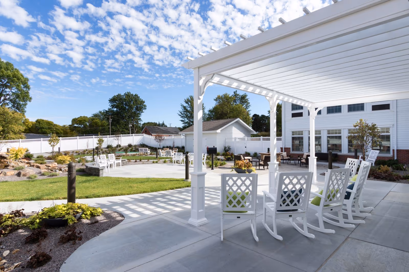 Outdoor patio area at CountryHouse Residence featuring white rocking chairs under a white pergola, a concrete walkway, green lawn, landscaped garden beds, and additional seating areas with chairs and tables. The background shows a white building with multiple windows and a white fence enclosing the area under a partly cloudy blue sky.